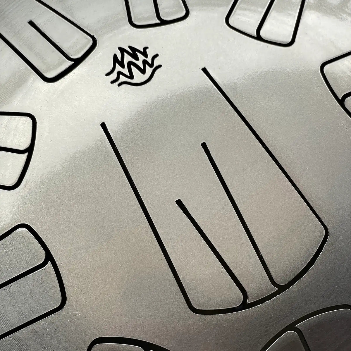 Close-up view of a metallic silver surface of a Spark V2 steel tongue drum with engraved flame-like Burning27 logo and petals.